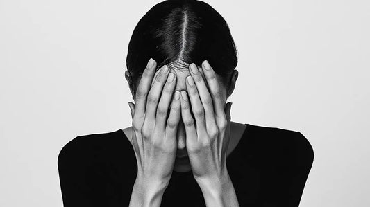 A black and white photograph of a woman with dark, sleek hair covering her face with her hands, expressing stress.