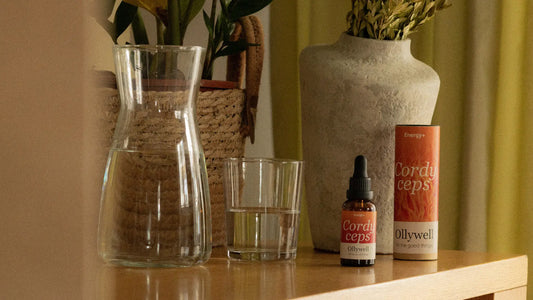 Cordyceps supplement in drops, set on a wooden surface next to a glass of water and a jug. In the background are potted plants and dried herbs in a vase.